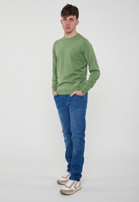 Maglietta verde a maniche lunghe con scollatura rotonda, abbinata a jeans blu e scarpe da ginnastica beige con dettagli bianchi. Design semplice, vestibilità casual.
