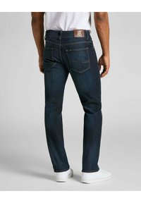 Jeans in denim blu scuro con un taglio slim, tasche posteriori visibili e un'etichetta in pelle marrone. Combinati con una sneaker bianca.