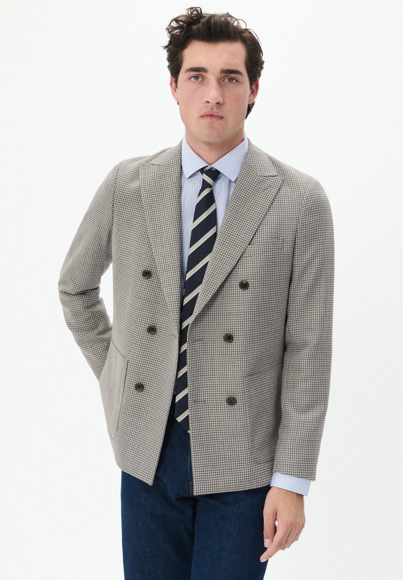 Kétgombos blézer houndstooth mintával, világosbarna és krémszínekben, sötét gombokkal. Kék csíkozott inggel és nyakkendővel párosítva.