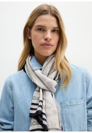 Frau mit langen blonden Haaren, die ein hellblaues Jeanshemd und einen grau gemusterten Schal um den Hals trägt.