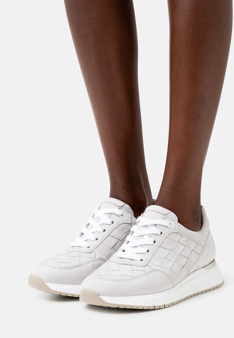 Personne portant des baskets en cuir tissé blanc avec des lacets blancs et des semelles beiges sur un fond clair.