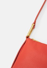 Rogaty torebka ze czerwonej skórki z charakterystyczną fakturą, z metalowymi złotymi akcentami i złączem paska na białym tle.