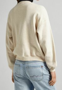 Beige sweatshirt met een geribde kraag en manchetten, gecombineerd met lichtblauwe jeans met een golvende stikselpatroon op de achterzakken.
