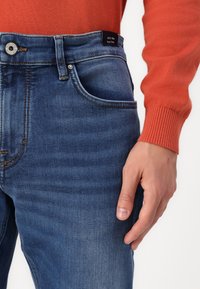 Calças de ganga azuis com desbotamento subtil, design de cinco bolsos, fecho de botão de metal e costura contrastante. Usadas com um suéter laranja.