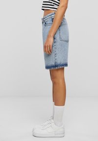 Person som har på seg en falmet blå dongeriskjørt i middels lengde, stripete crop top, hvite sokker og hvite joggesko, står i profil på en grå bakgrunn.