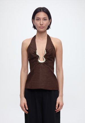 Žena s krátkymi tmavými vlasmi, ktorá má na sebe zlaté kruhové náušnice, hnedý top s ramienkami a zlatým kovovým zapínaním uprostred a čierne nohavice, stojaca na bielom pozadí.