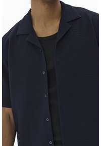 Camicia a maniche corte in tessuto navy strutturato, con chiusura a bottoni e colletto a punta sopra una canotta nera.