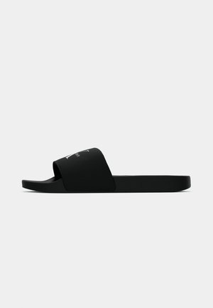 Black slide sandal with wide fabric strap featuring white Calvin Klein logo, thick sole, and open toe, viewed from the side on white background.