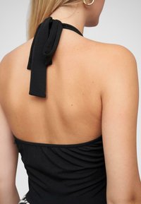 Haut halter noir avec une bride nouée dans le dos, un design en tissu froncé et une texture lisse sans embellissements supplémentaires.