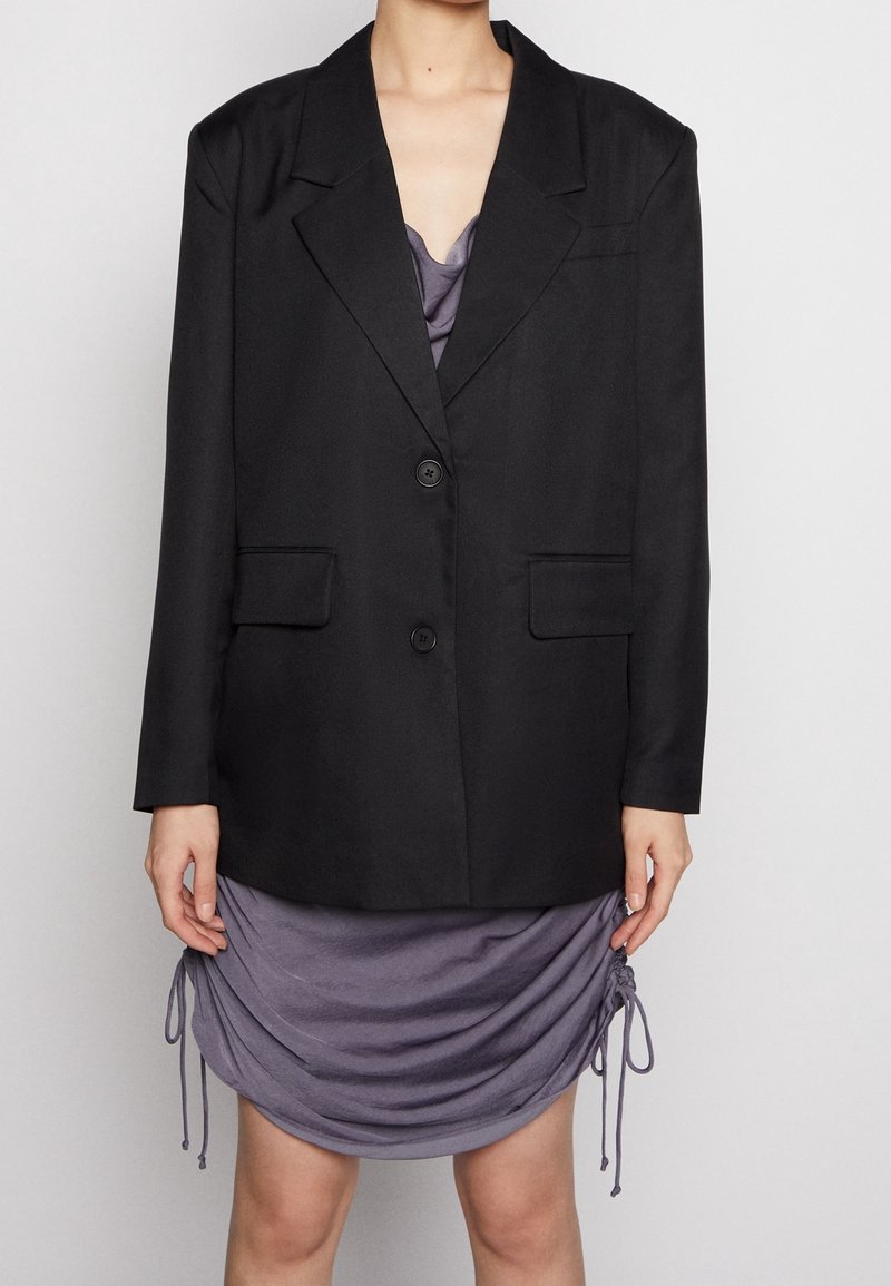 Svart kavaj med två knappar, spetsiga slag och två framfickor, lager över en draperad lila klänning med dragsnodd detaljer.