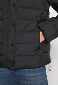 Black puffer jacket, quilted design, smooth texture, streamlined silhouette, featuring snap buttons and side pocket with adjustable drawstring.