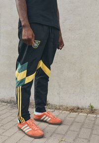 Pantalones deportivos negros con patrones en forma de chevron en verde y amarillo, acentuados con tres rayas amarillas. Zapatillas de deporte brillantes en naranja con rayas en azul verdoso.