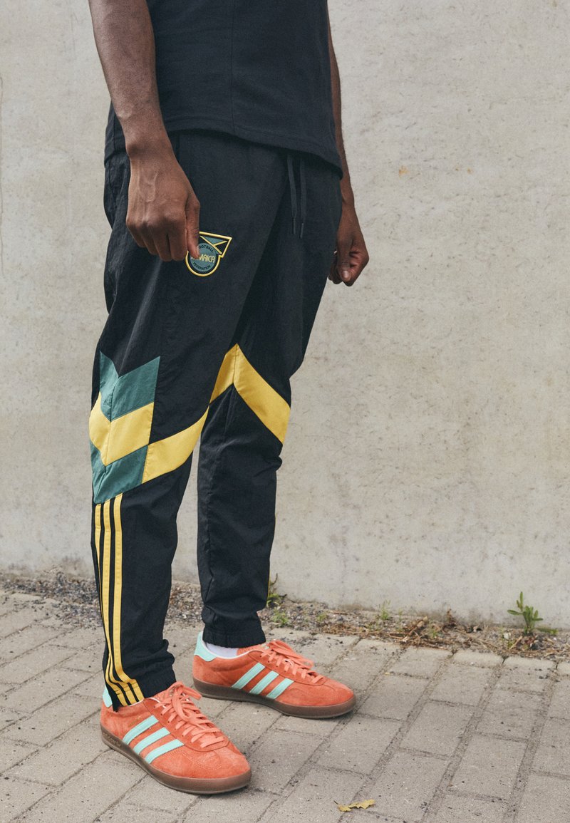 Pantalones deportivos negros con patrones en forma de chevron en verde y amarillo, acentuados con tres rayas amarillas. Zapatillas de deporte brillantes en naranja con rayas en azul verdoso.