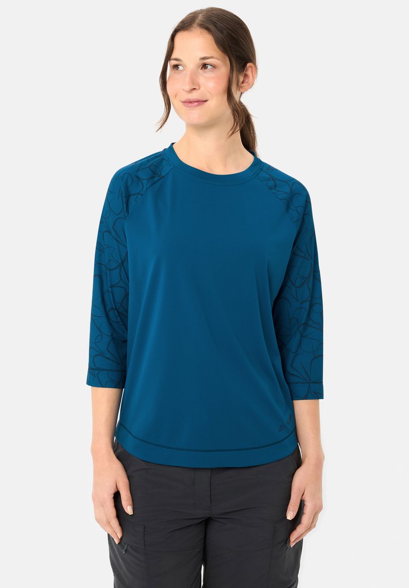 Jeune femme portant un t-shirt à manches longues turquoise avec des manches sombres à motifs et un pantalon noir, debout devant un fond clair uni.
