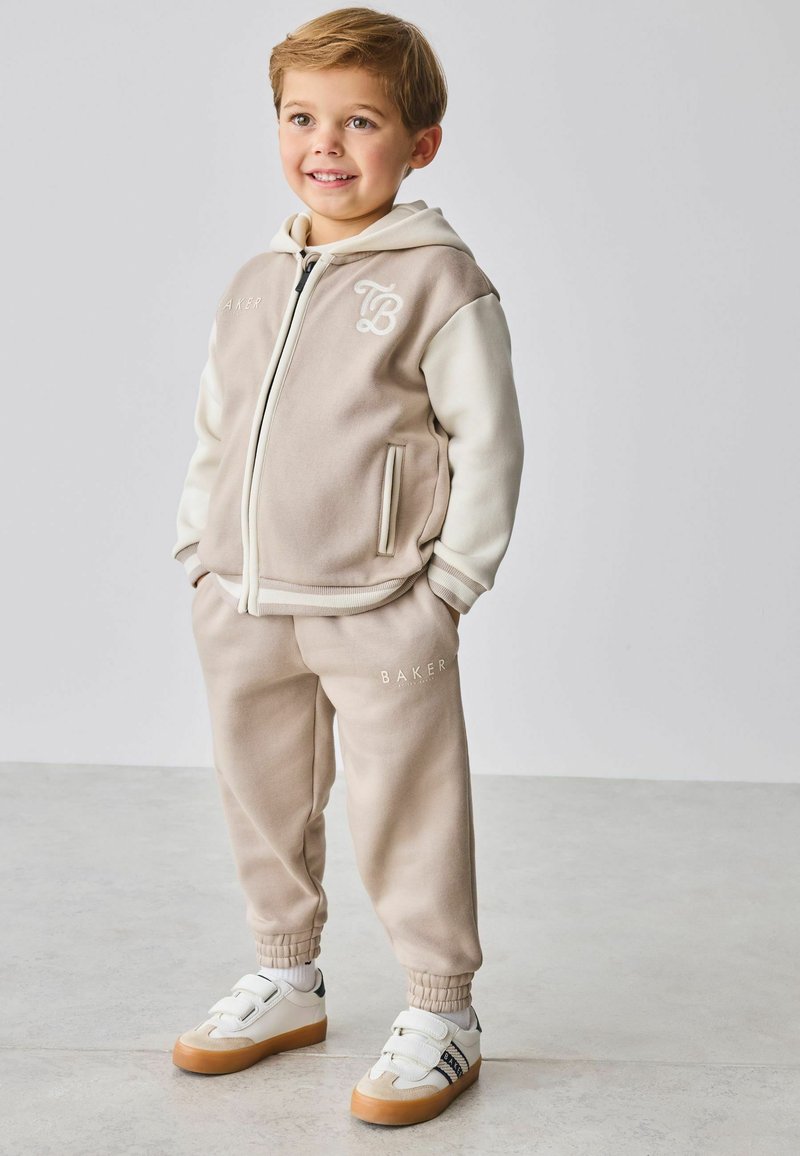 Beżowy i kremowy dres z bluzą zapinaną na zamek i spodniami, z prążkowanymi mankietami i logo „BAKER”. Dopełniony białymi sneakersami z gumową podeszwą.