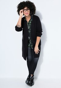 Femme aux cheveux bouclés portant des lunettes, une chemise à motifs, un long cardigan noir, un jean foncé et des bottines noires, posant contre un mur blanc.