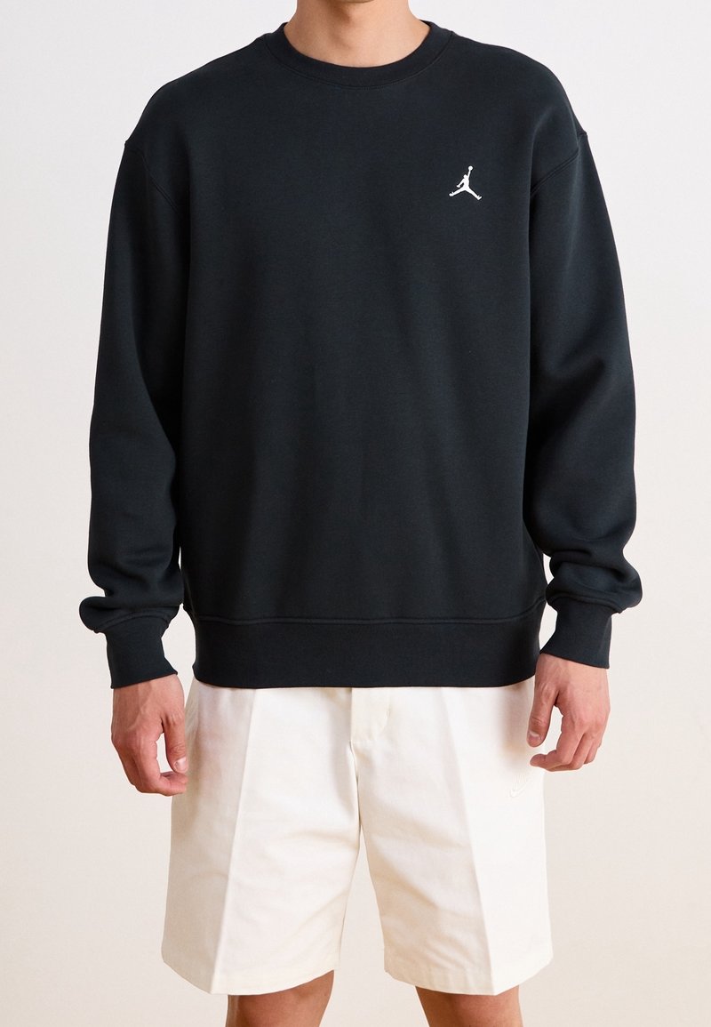 Homme portant un sweat-shirt noir à col rond avec un logo Jumpman blanc et un short blanc jusqu'aux genoux.