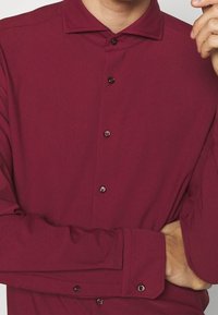 Camisa de manga larga color marrón burdeos con cuello de camisa abotonado, que presenta botones oscuros y una textura suave. Los puños son ligeramente ajustados y tienen detalles de botones.