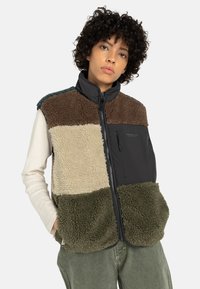 Model draagt een kleurgeblokt, fleec gevoerd vest met tinten bruin, crème en groen, gecombineerd met een langhands T-shirt en relaxte broek.