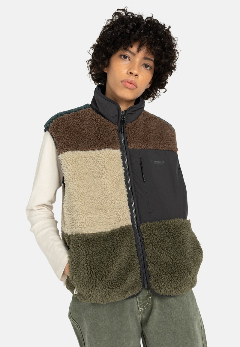 Model draagt een kleurgeblokt, fleec gevoerd vest met tinten bruin, crème en groen, gecombineerd met een langhands T-shirt en relaxte broek.