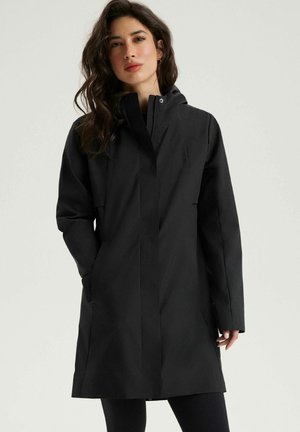 Kobieta o długich ciemnych włosach, ubrana w czarny płaszcz sięgający do kolan z wysokim kołnierzem, z rękami w kieszeniach, stojąca na jednolitym jasnym tle.