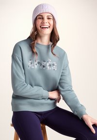 Light blue sweatshirt with floral "Après" text, crew neck, and ribbed cuffs. Paired with dark purple leggings. Woolen light purple beanie.