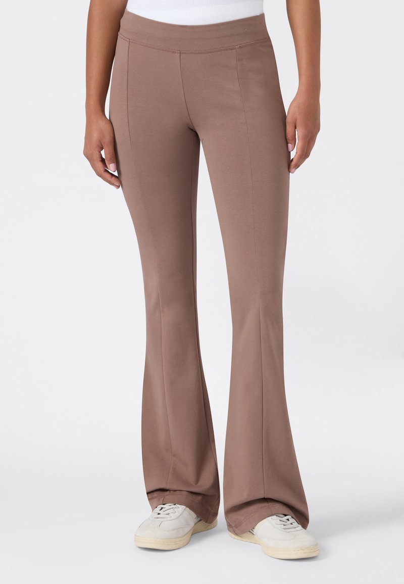 Personne portant un pantalon flare taille haute marron taupe et des baskets blanches, debout devant un fond uni.