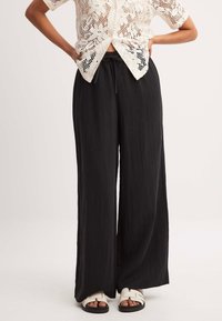 Zwarte wijde broek van lichtgewicht, gestructureerde stof met een elastische tailleband en trekkoord. Gecombineerd met een crèmekleurige kanten top en witte sandalen.
