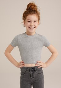 Grå ribbat kortärmad croptop som kombineras med högmidjade mörka denimjeans. Modellen poserar med händerna på höfterna, leende mot en neutral bakgrund.