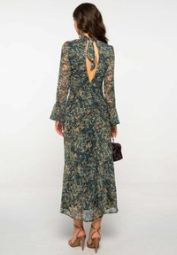 Mujer con cabello largo y ondulado lleva un vestido de manga larga, hasta la pantorrilla, con un estampado en verde y marrón, con un corte en la espalda y un lazo, sosteniendo un pequeño bolso oscuro.