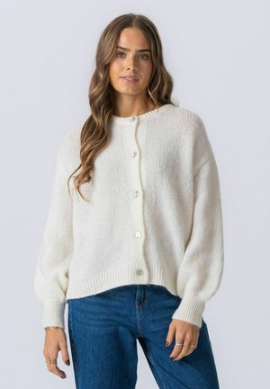 Žena s dlhými vlnitými vlasmi, oblečená v bielom kardigáne na gombíky a modrých džínsach, stojaca pred jednoduchým svetlým pozadím.