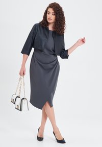 Abito grigio con design drappeggiato e orlo asimmetrico, abbinato a una giacca nera cropped, tacchi alti neri e una borsa bianca e nera.