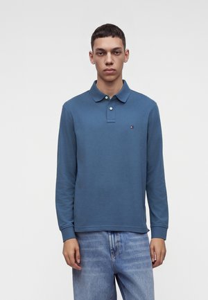 Jeune homme portant un polo bleu à manches longues et un jean bleu clair, debout devant un fond clair uni.