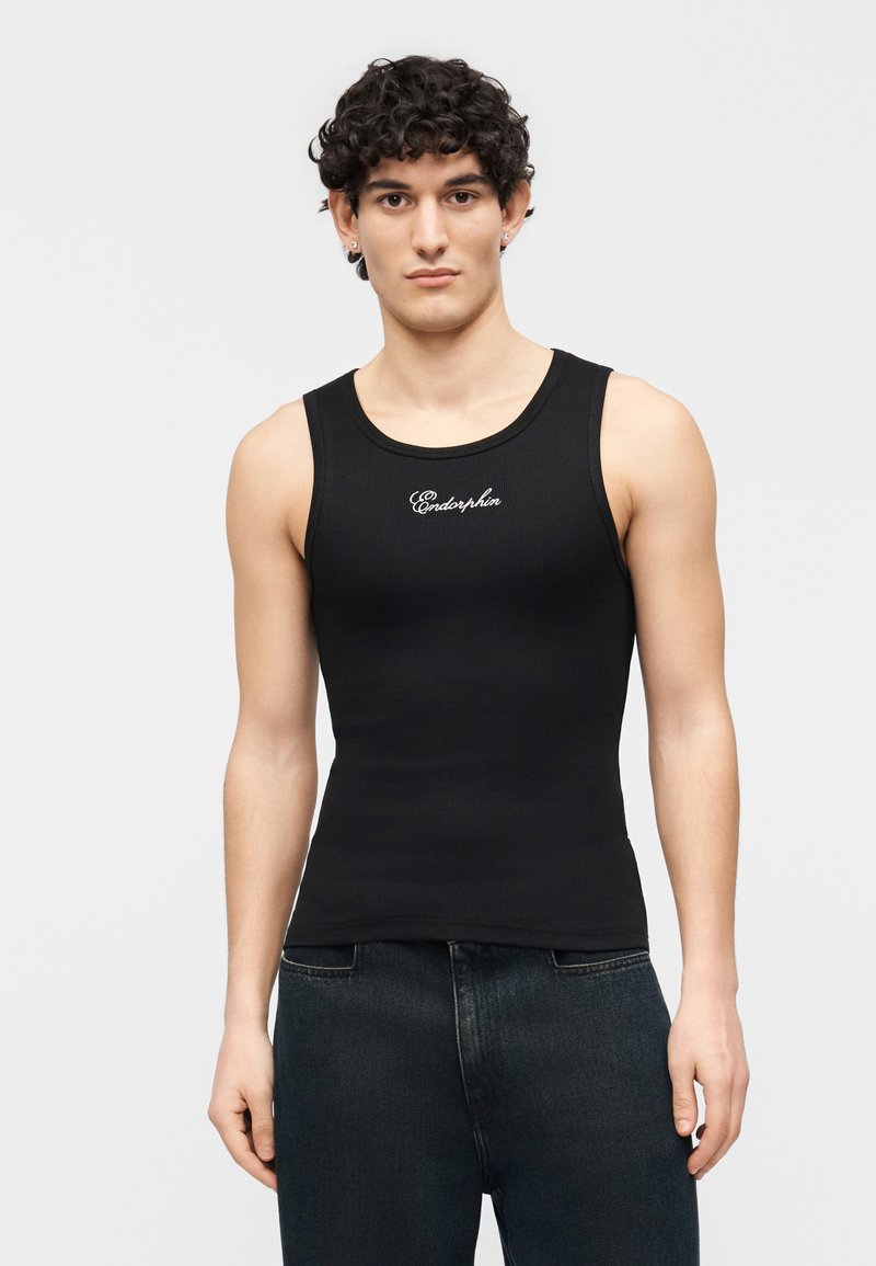 Jeune homme aux cheveux bouclés portant un haut noir sans manches côtelé avec le texte "Endorphin", associé à un jean foncé, debout devant un fond uni.