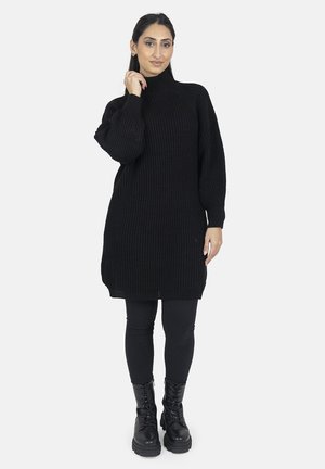 Robe pull noire en tricot oversize avec un col haut, des manches longues et une texture côtelée, associée à des leggings noirs et des bottes à lacets.