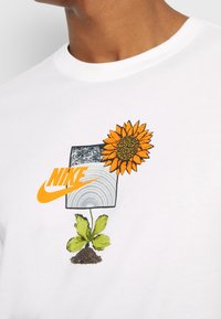 Camiseta blanca de algodón con gráficos coloridos: un girasol, una planta brotando y el logo de Nike en naranja sobre un fondo cuadrado gris.