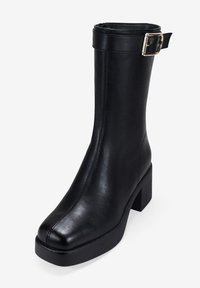 Botín negro de cuero hasta el tobillo con puntera cuadrada, tacón grueso y un detalle de hebilla en la correa en la parte superior. Textura suave y diseño minimalista.