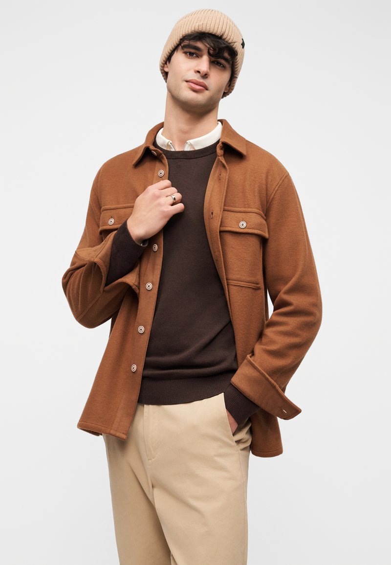 Junger Mann mit beiger Mütze, brauner Jacke mit Knöpfen über einem dunkelbraunen Pullover und beiger Hose, stehend mit einer Hand in der Tasche.