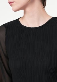 Črn top z nabranim teksturo in okroglim izrezom, z prozornimi črnimi rokavi. Gladka tkanina z rahlim sijajem, minimalističen dizajn.