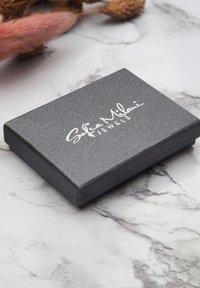 Grå tekstureret smykkeskrin med fladt design, der har et sølvlogo på toppen. Overfladen har en subtil glans.