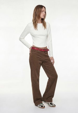 Ung kvinde iført hvid langærmet top, brune bukser med vide ben og rød bælte, samt hvide sko med sorte og grønne detaljer, stående foran en hvid baggrund.