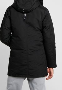 Zwarte geïsoleerde parka met een capuchon, voorzien van een ritssluiting, zijzakken en een gladde textuur. Het ontwerp is knie-lengte met een rechte snit.