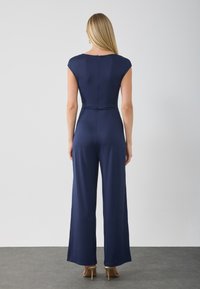 Mörkblå jumpsuit med ärmveck, figursydd överdel, utsvängda byxor och en dragkedja på ryggen. Slätt tyg med en mjuk textur.
