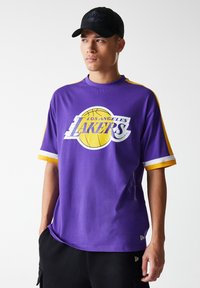 Lila t-shirt med kort ärm och gula detaljer, med en vit Los Angeles Lakers-logotyp och en basketgrafik. Tillverkad av bomull.