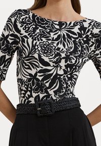 Woman wearing black and white floral patterned top with elbow-length sleeves and a black braided belt over black pants.