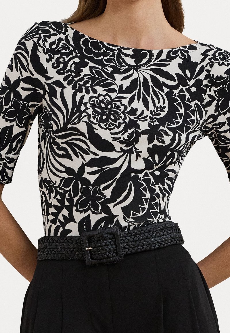 Woman wearing black and white floral patterned top with elbow-length sleeves and a black braided belt over black pants.