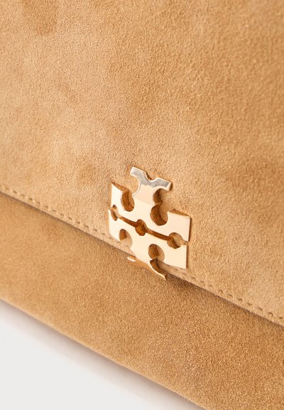 Λεπτομέρεια τσάντας ώμου σε απόχρωση ταμπά σουέντ με χρυσό μεταλλικό έμβλημα λογότυπου κεντραρισμένο κοντά στο κάτω άκρο του καπακιού.