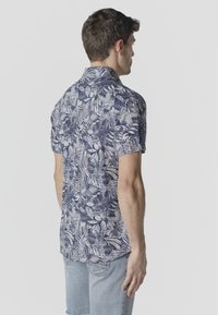 Homme portant une chemise à manches courtes à motif tropical bleu marine et blanc, vu de dos, associée à un short en denim bleu clair.