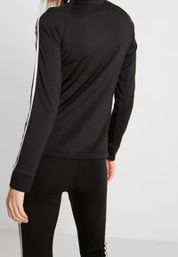 Osoba v černém sportovním oblečení s bílými pruhy na rukávech a bocích, stojící proti jednoduchému bílému pozadí.