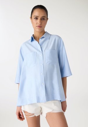 Donna che indossa una camicia azzurra chiara, larga, con bottoni e due tasche sul petto, e pantaloncini bianchi consumati, su uno sfondo semplice.